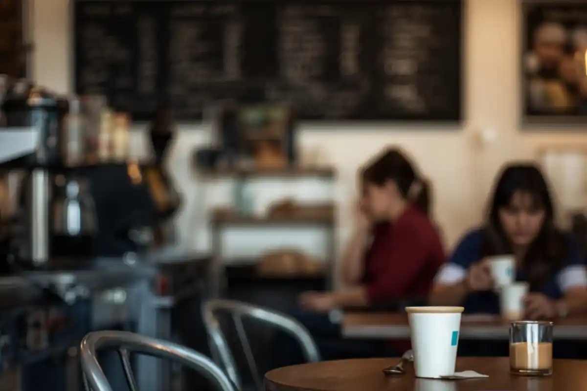 Starbucks kavárna s rozostřeným pozadím, v popředí papírový kelímek s kávou na stole, zatímco návštěvníci sedí uvnitř podniku.